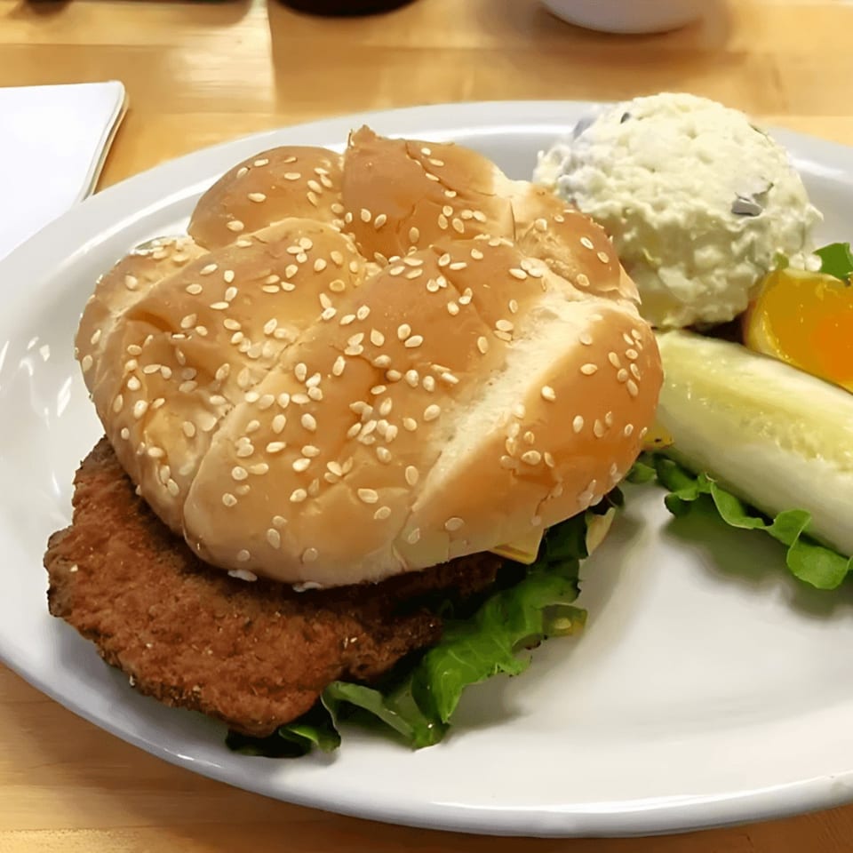 A breaded pork tenderloin sandwich with lettuce, a pickle spear, and potato salad on a white plate.