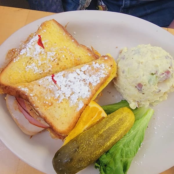 A Monte Cristo sandwich with powdered sugar, potato salad, a pickle, orange slice, and lettuce.